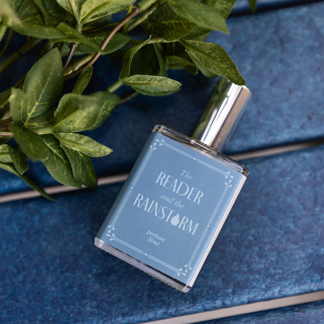 A close-up image of the bottle of perfume scented "The Reader and the Rainstorm" showing the light and dark blues of the bottle's details. The bottle lays on a blue slatted background with greenery at the top of the image.