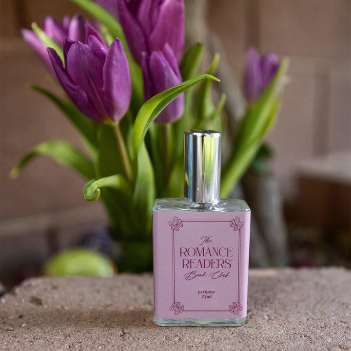 A close-up image of the bottle of perfume scented "The Romance Readers' Book Club" showing the light and dark pink details on the bottle label. The bottle stands with a bouquet of purple tulips in the background.