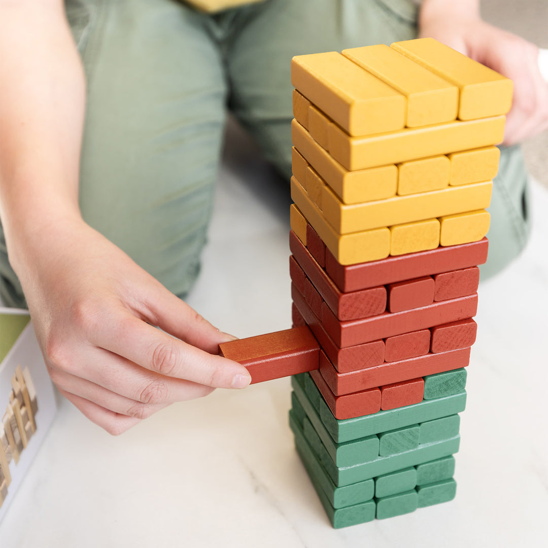 A pair of white hands pulls out a red block from a multi-colored brick tower in the tumbling brick game.