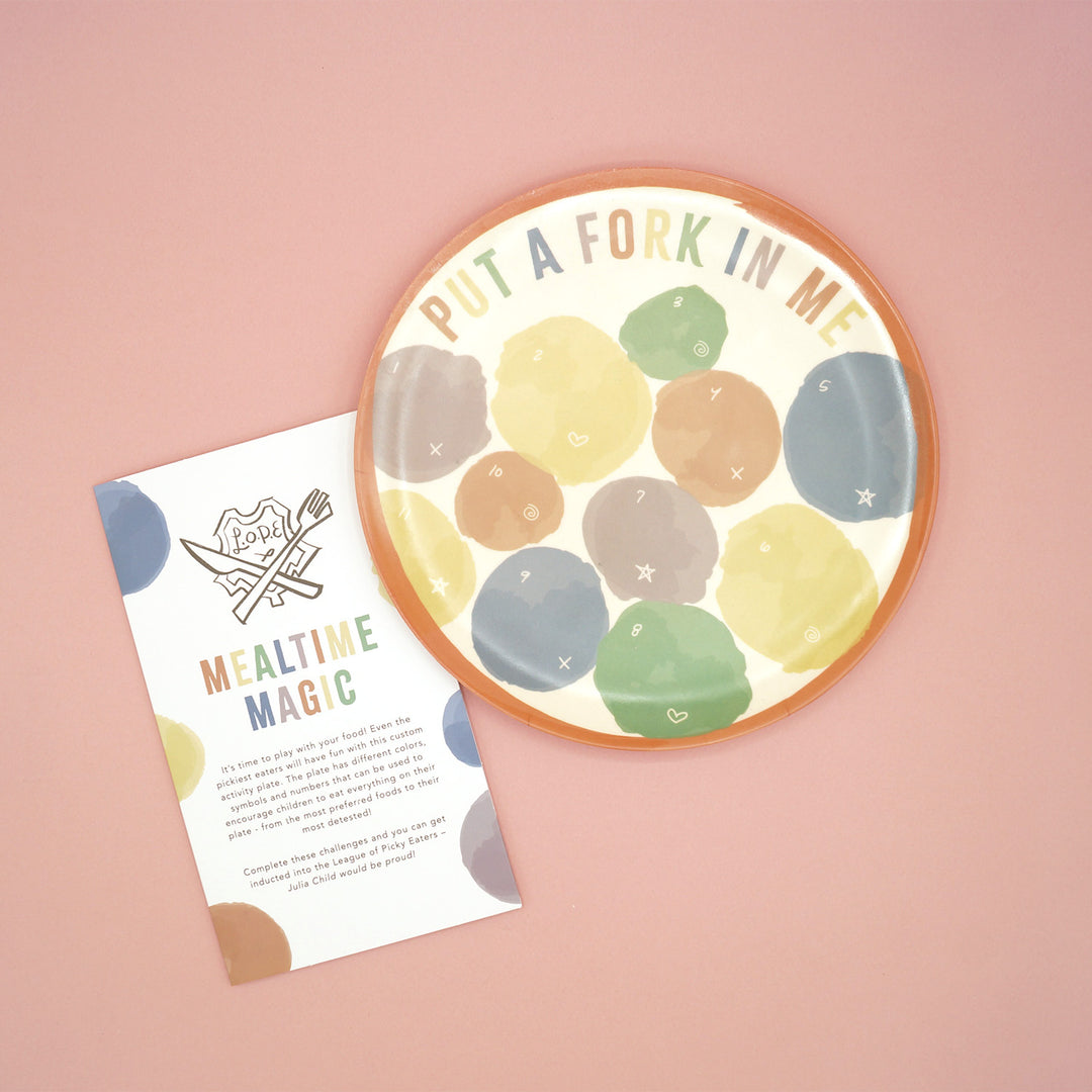 A multi-colored plate sits on a pink background. The plate says "Put a Fork in Me" along the top and has multiple colored dots on the plate labeled with numbers and symbols for various games. The instruction booklet sits next to the plate in the image. 