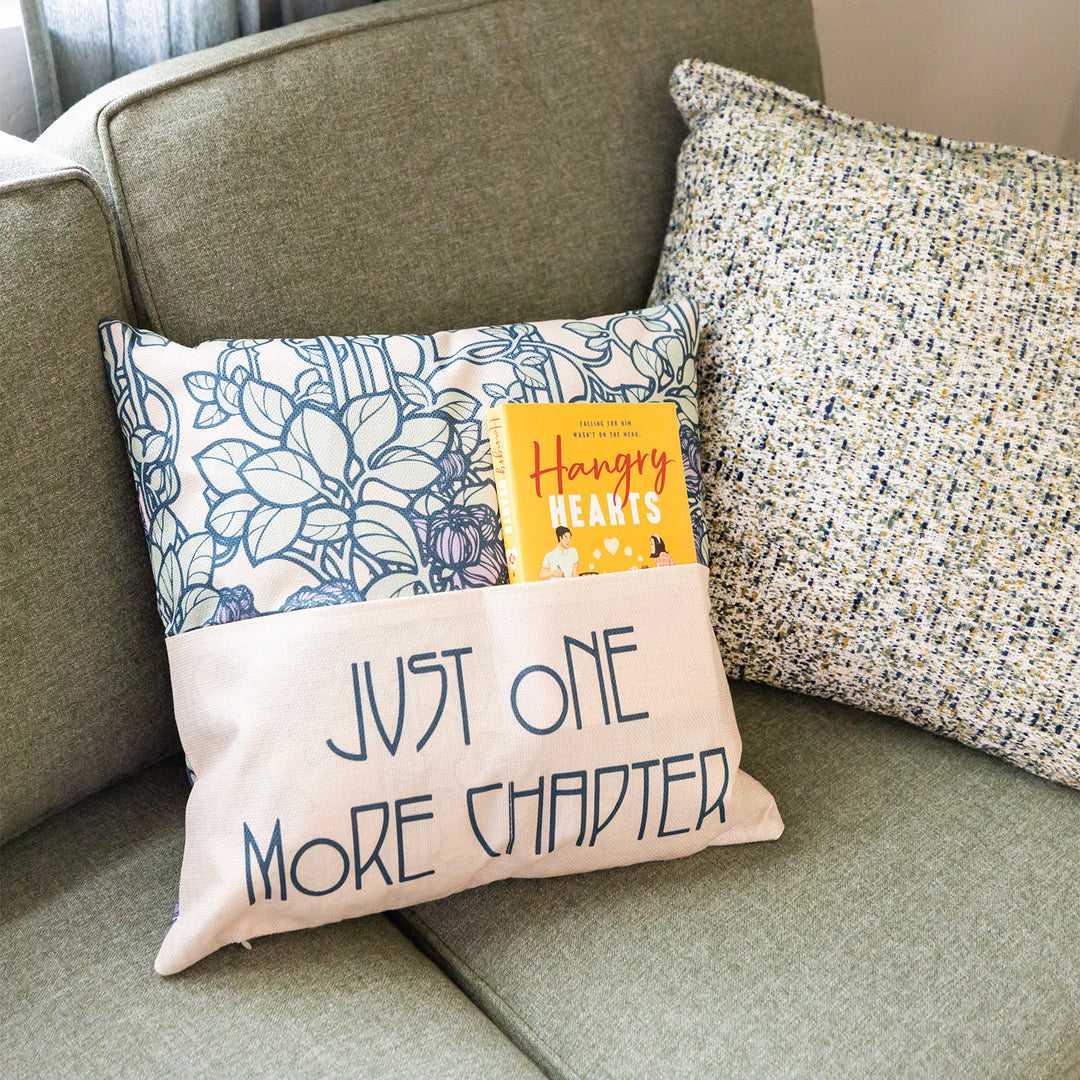 A square light tan pillow with light green/purple floral pattern lays on a couch. The front of the pillow has a pocket that covers the bottom half, with the words Just One More Chapter across it. A paperback edition of Hangry Hearts is tucked into the pocket.