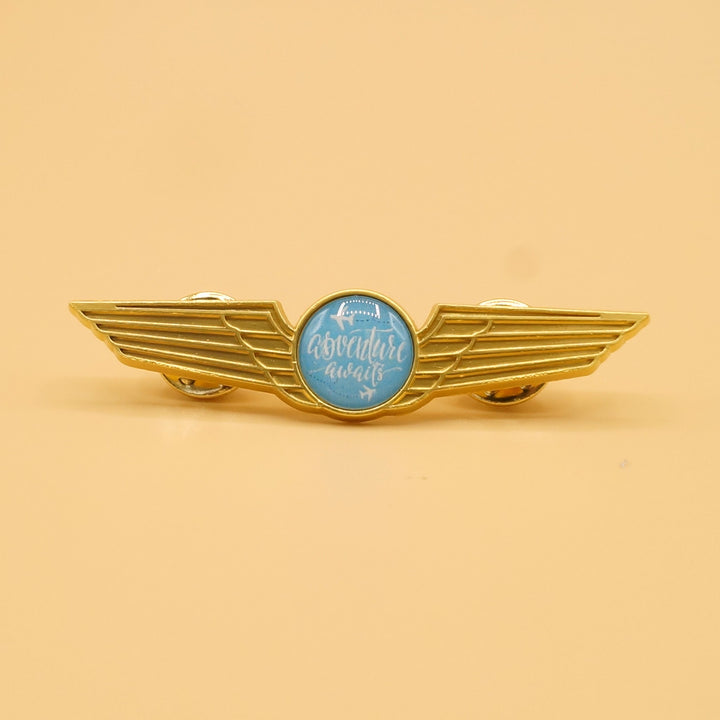 an enamel pin with gold wings and a blue circle at the center that says "Adventure Awaits"