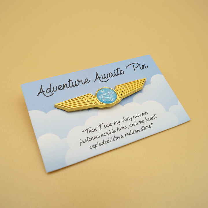 an enamel pin with gold wings and a blue circle at the center that says "Adventure Awaits" sits on a square with a pattern of clouds