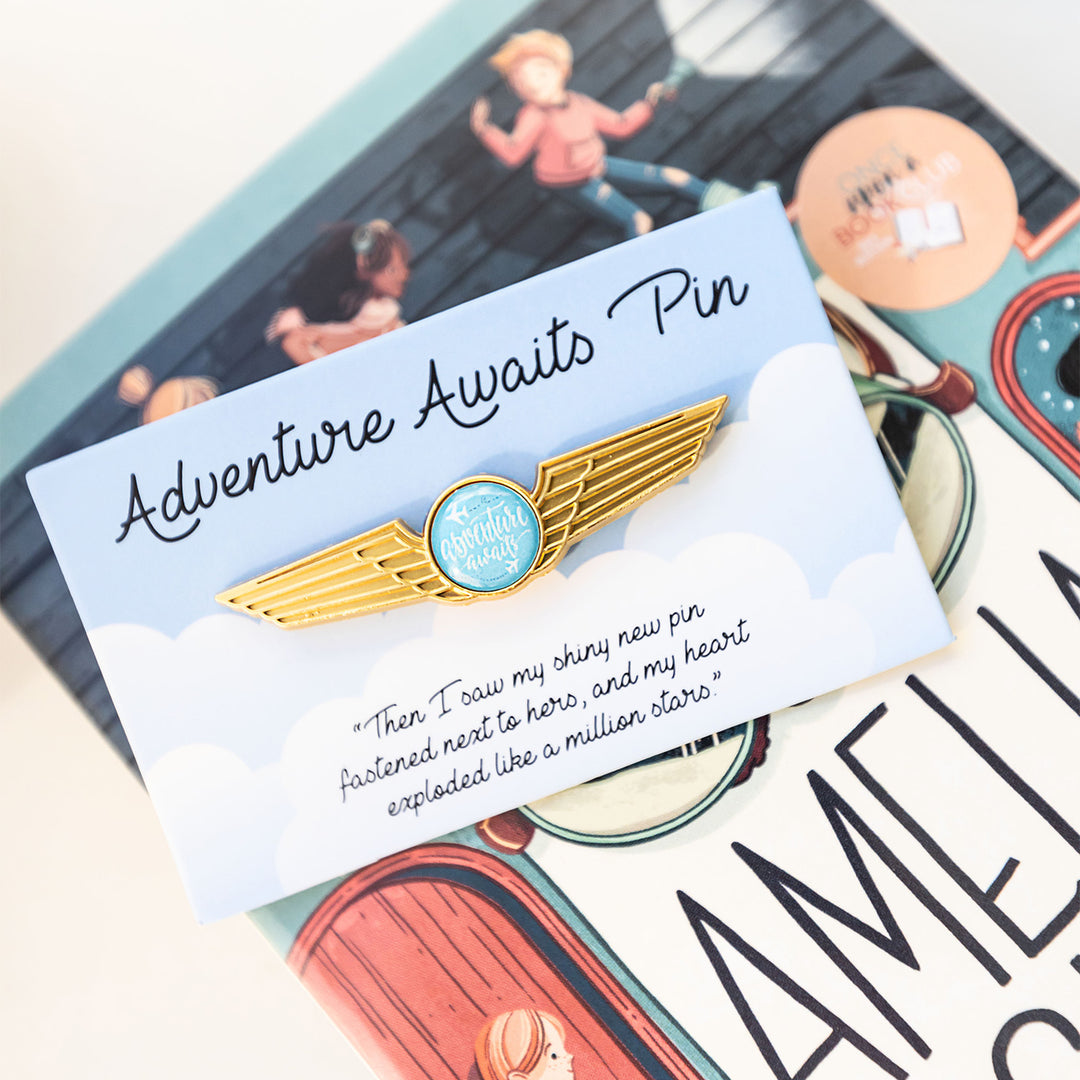 an enamel pin with gold wings and a blue circle at the center that says "Adventure Awaits" sits on a square with a cloud pattern. Sitting against a paperback copy of The Amelia Six