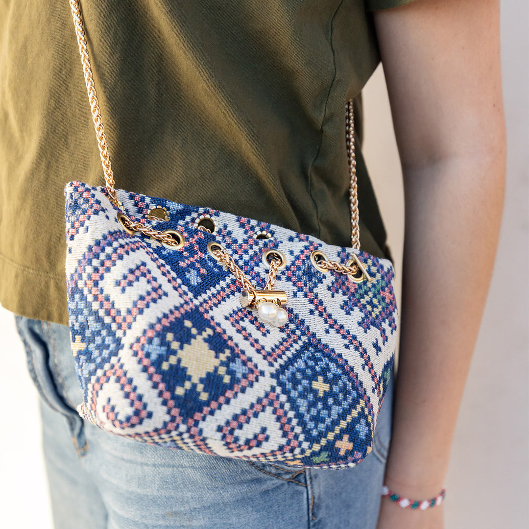 A round bubble purse with a woven, geometric pattern and gold chain strap is worn by a woman wearing jeans and a green shirt. Purse is closed with a gold chain slider clasp with faux-pearl details.