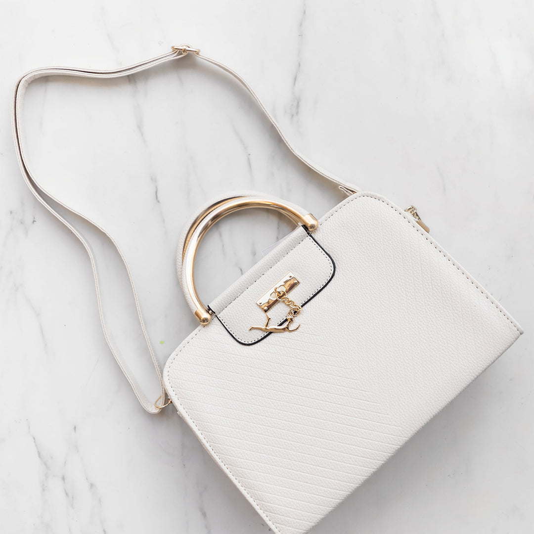 A white marble floor. A white rectangular satchel purse lays on the marble flooring. A gold charm hangs from the front. A long shoulder strap is stretched to show length.