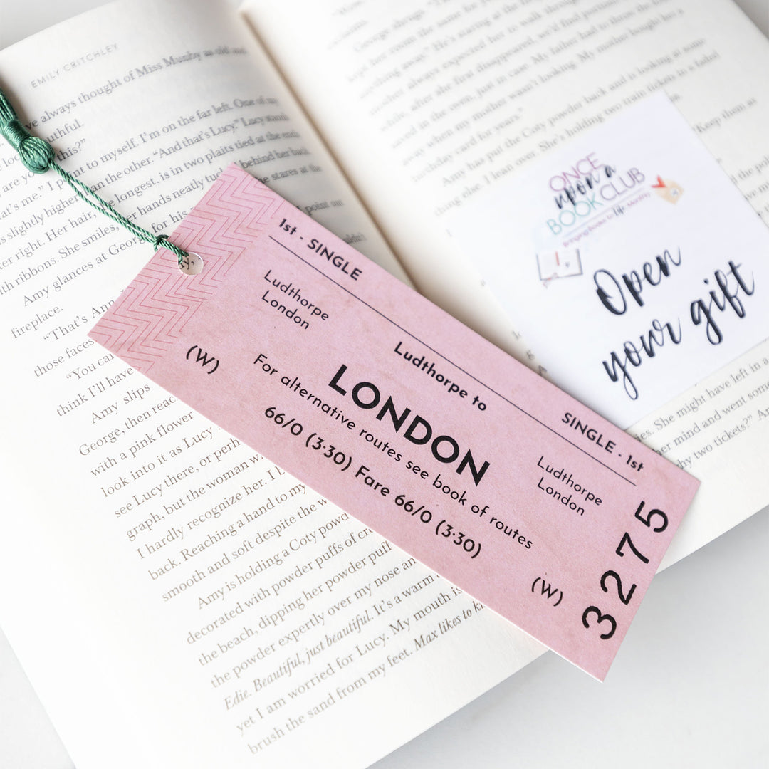 A pink bookmark designed to look like a train ticket from Ludthorpe to London with a green tassel. Sits on an open book with an Open Your Gift sticky note visible behind it.