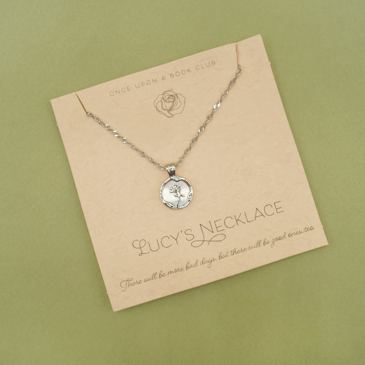 A small silver pendant with a rose sits on necklace card. The words "Lucy's necklace" is written along the bottom of the card.