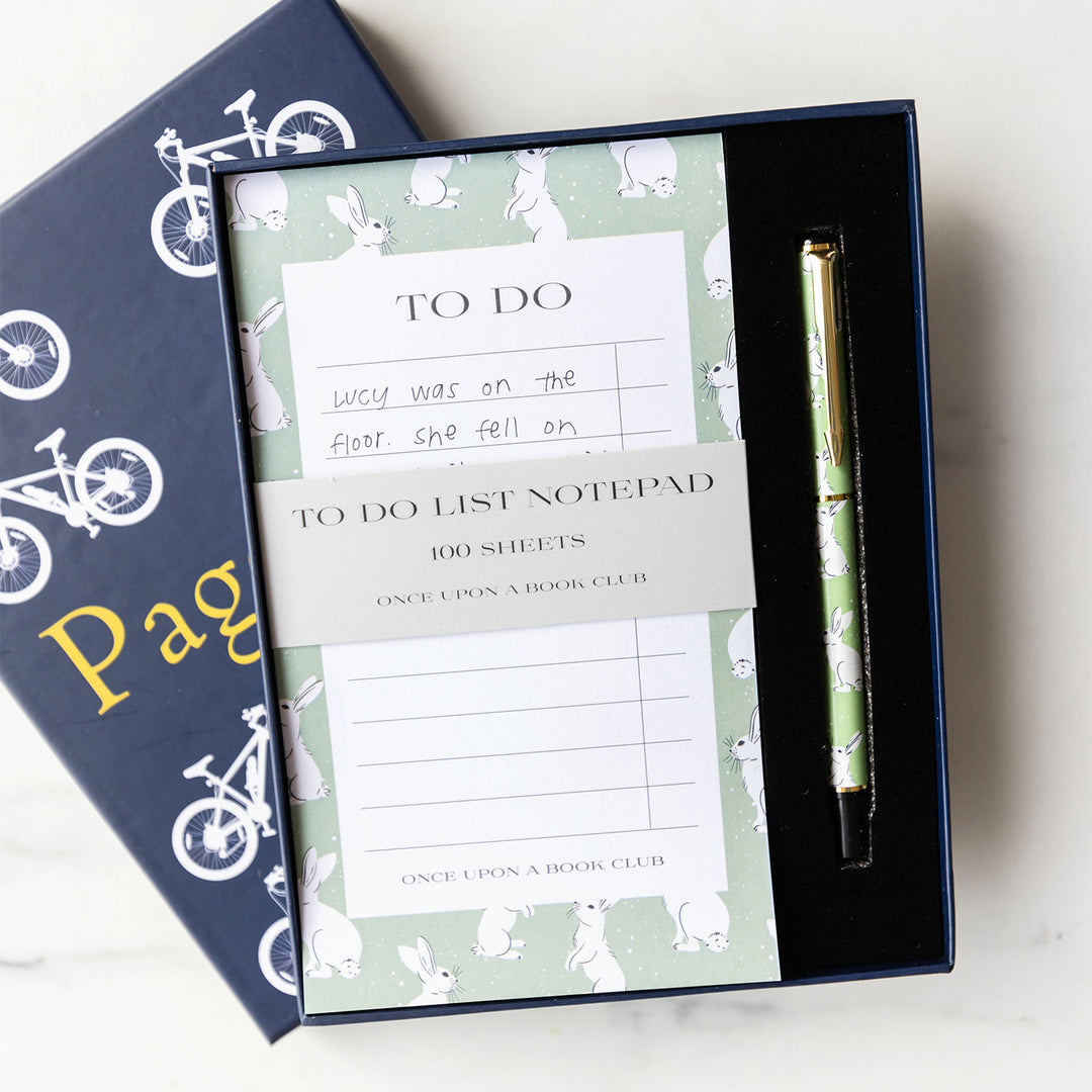 An open box shows a light green to do list patterned with rabbits around the border. The paper wrapping around the notepad shows that it is 100 sheets. The pen sitting next to the note pad in the foam within the box is also patterned green with white rabbits and gold detailing.