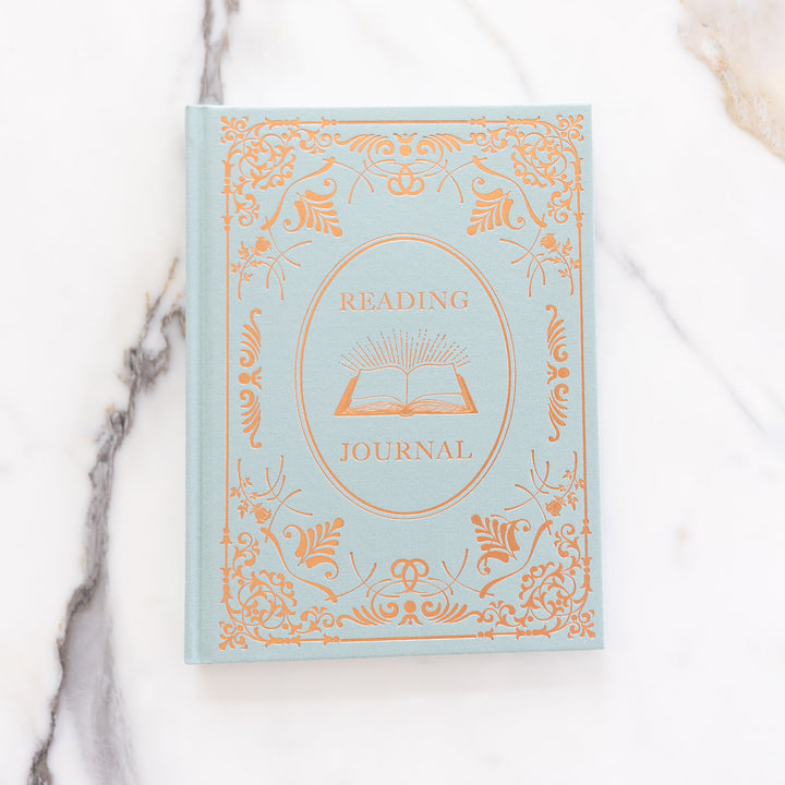 A flat lay of the seafoam blue and bronze reading journal on a marble countertop.