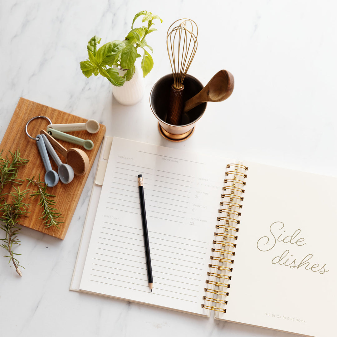 A recipe book lays open, the left page is lined with a pencil laying across the page and the right page says Side dishes. Above the recipe book are a cup with a whisk and wooden spoon in it, a set of measuring spoons, and a plant