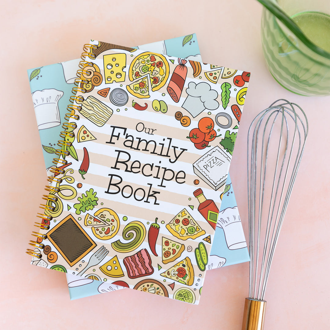 A spiral bound notebook with images of food surrounding the words Our Family Recipe Book. The book is laying on a blue box with a chefs hat pattern, next to a whisk