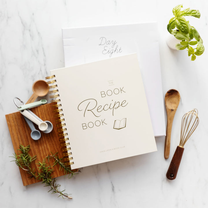 A white Book Recipe Book is centered, surrounded by a set of measuring spoons, a wooden spoon, a whisk, and a plant. A piece of paper labeled Day Eight is sticking out of the recipe book
