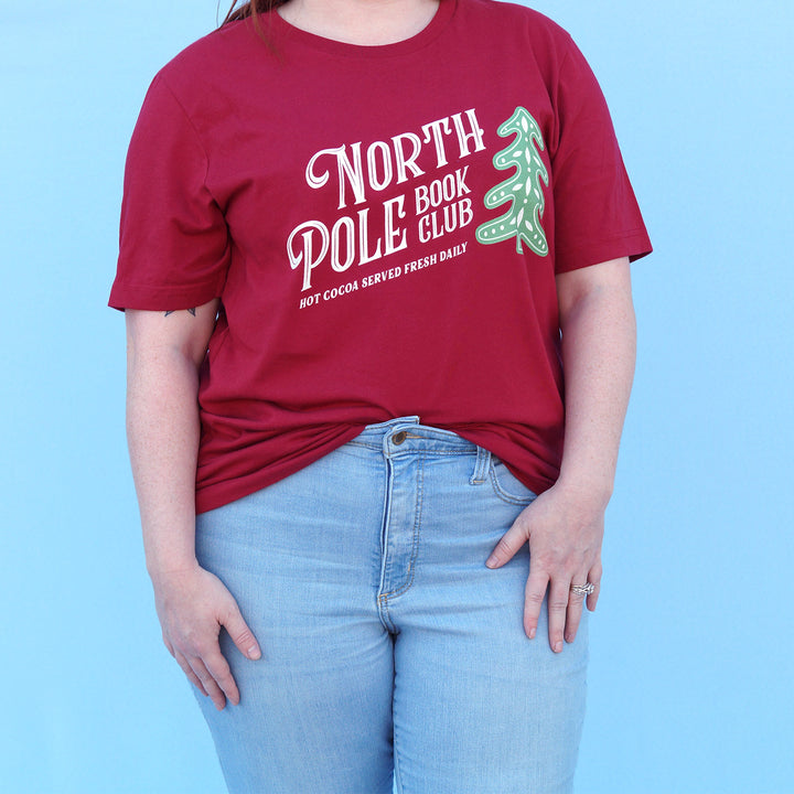 Person wearing a red t-shirt with 'North Pole Book Club' text and graphic on a blue background