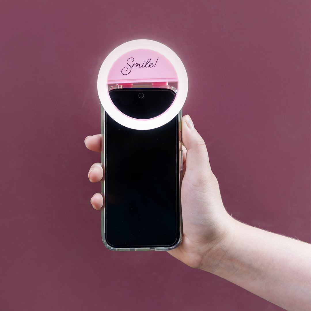 A pink ring light that says Smile! attached to the top of an iPhone held out in front of a purple background