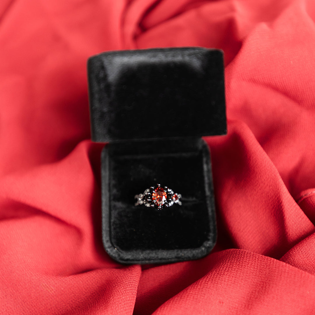 A black ring with a red gemstone in the center sits in a black ring box on red fabric