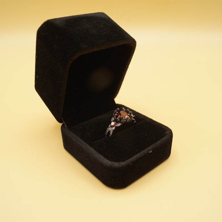 a black ring with a red gemstone sits in a black ring box against a yellow background