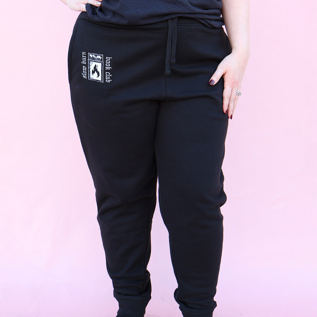 Black sweatpants with embroidered 'Slow Burn Book Club' and matchbook design on a pink background
