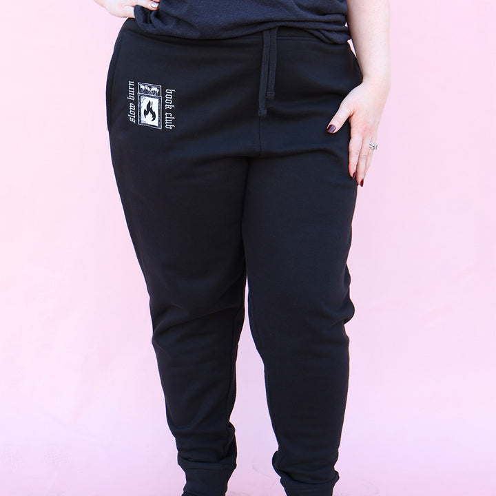 Black sweatpants with embroidered 'Slow Burn Book Club' and matchbook design on a pink background