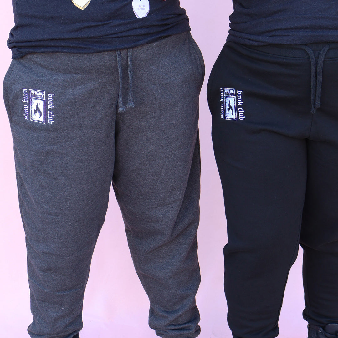 Gray and black sweatpants with embroidered 'Slow Burn Book Club' and matchbook design on a pink background