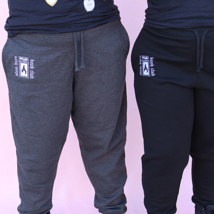 Gray and black sweatpants with embroidered 'Slow Burn Book Club' and matchbook design on a pink background
