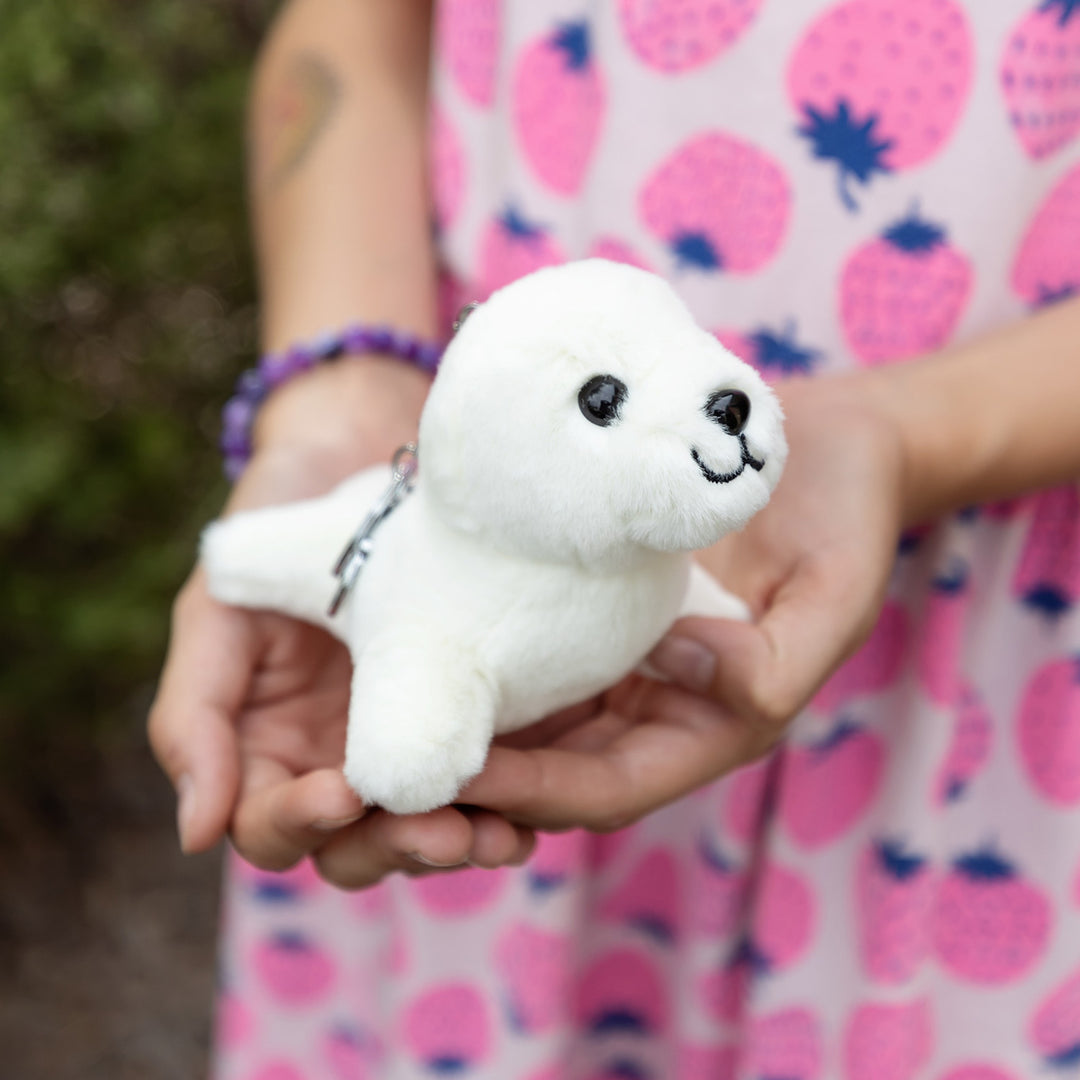 A white plush seal on a keychain is held in two hands in front of a girl wearing a strawberry-patterned dress.