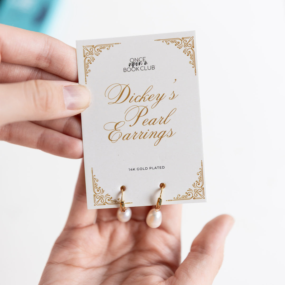 Hands hold up a card that says Once Upon a Book Club Dickey's Pearl Earrings. A pair of gold, pearl earrings hang from the card.