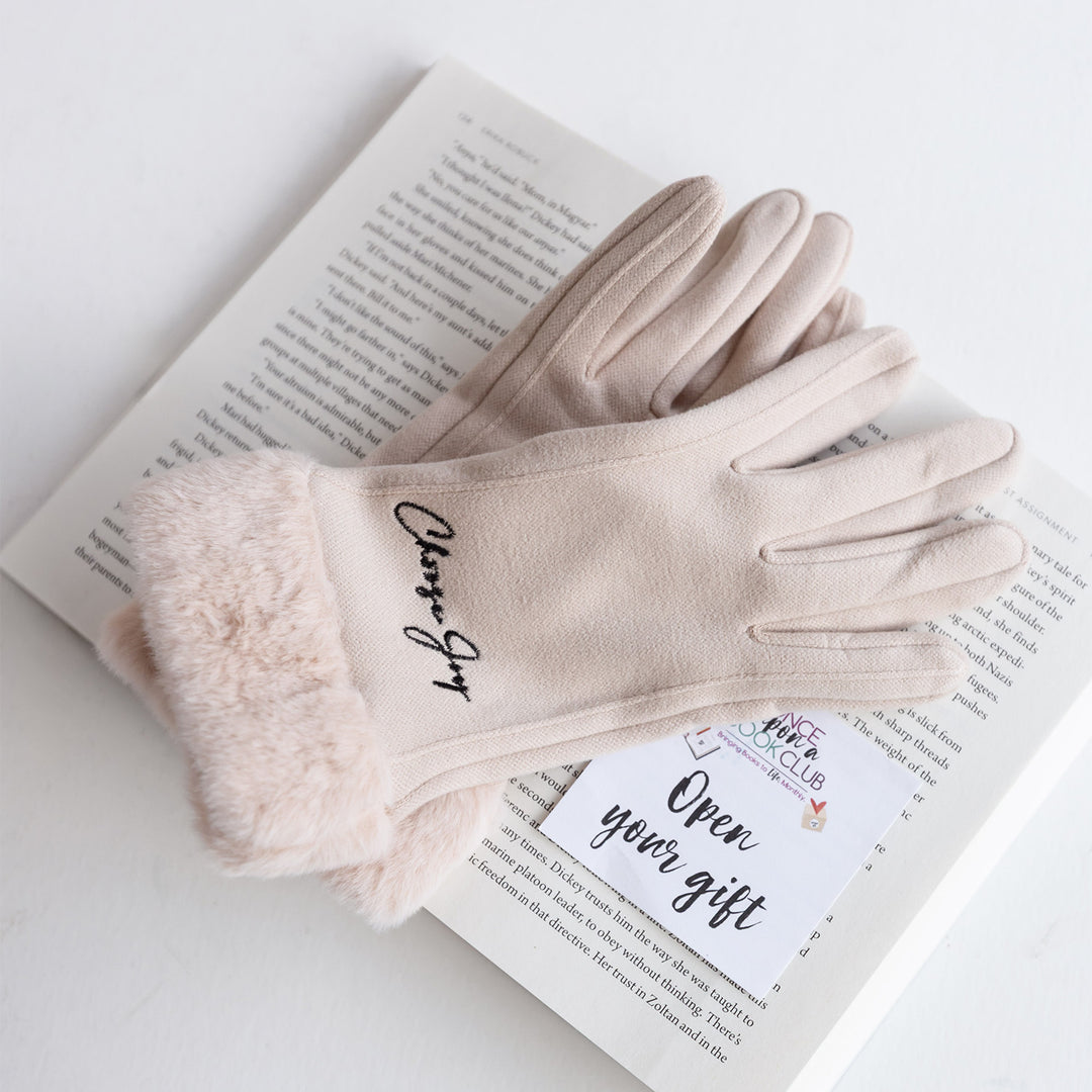 A pair of light pink faux fur winter gloves laying on an open book. On the back of the glove, black text says Choose Joy.