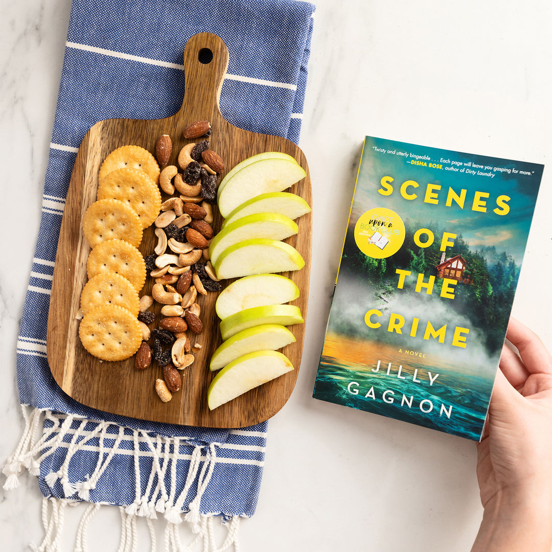 a white hand holds a paperback copy of "Scenes of the Crime" next to a wooden serving board with apple slices, nuts, and ritz crackers on it, laying on a blue towel.