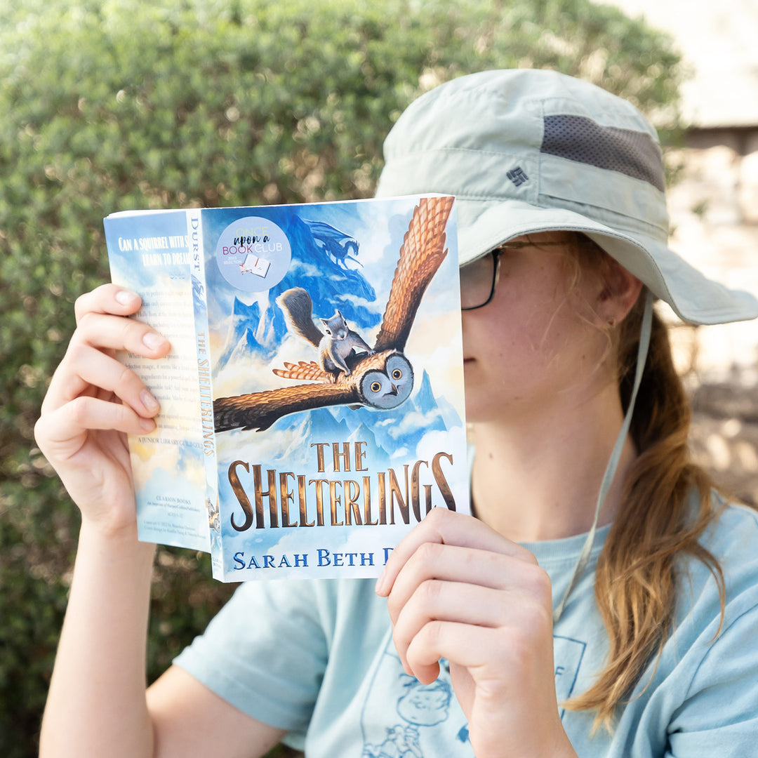 A child holds an open copy of The Shelterlings by Sarah Beth Durst outdoors and reads intently.