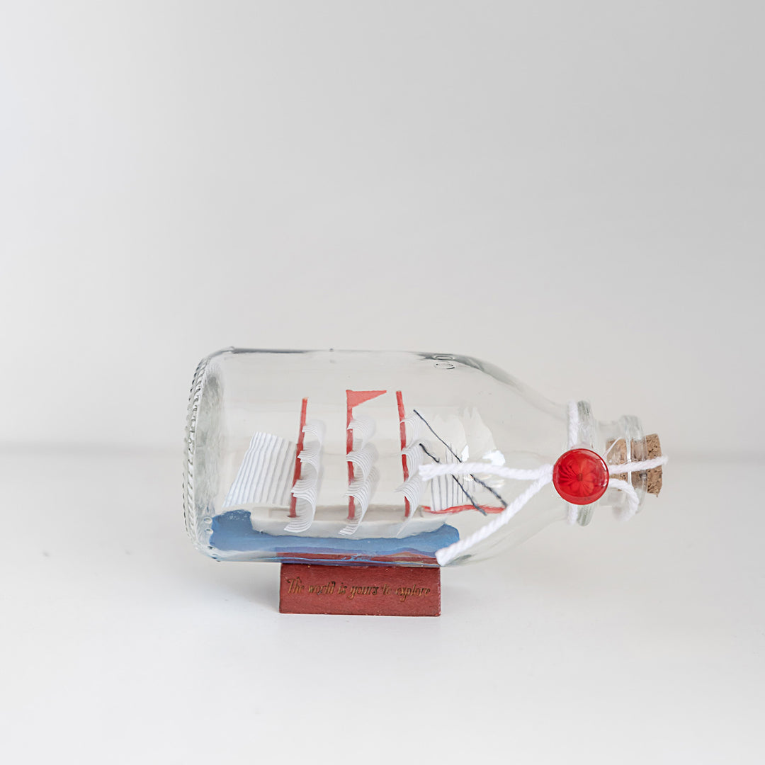 A glass bottle with a small ship in it and the words The World is Yours to Explore on the wooden base.