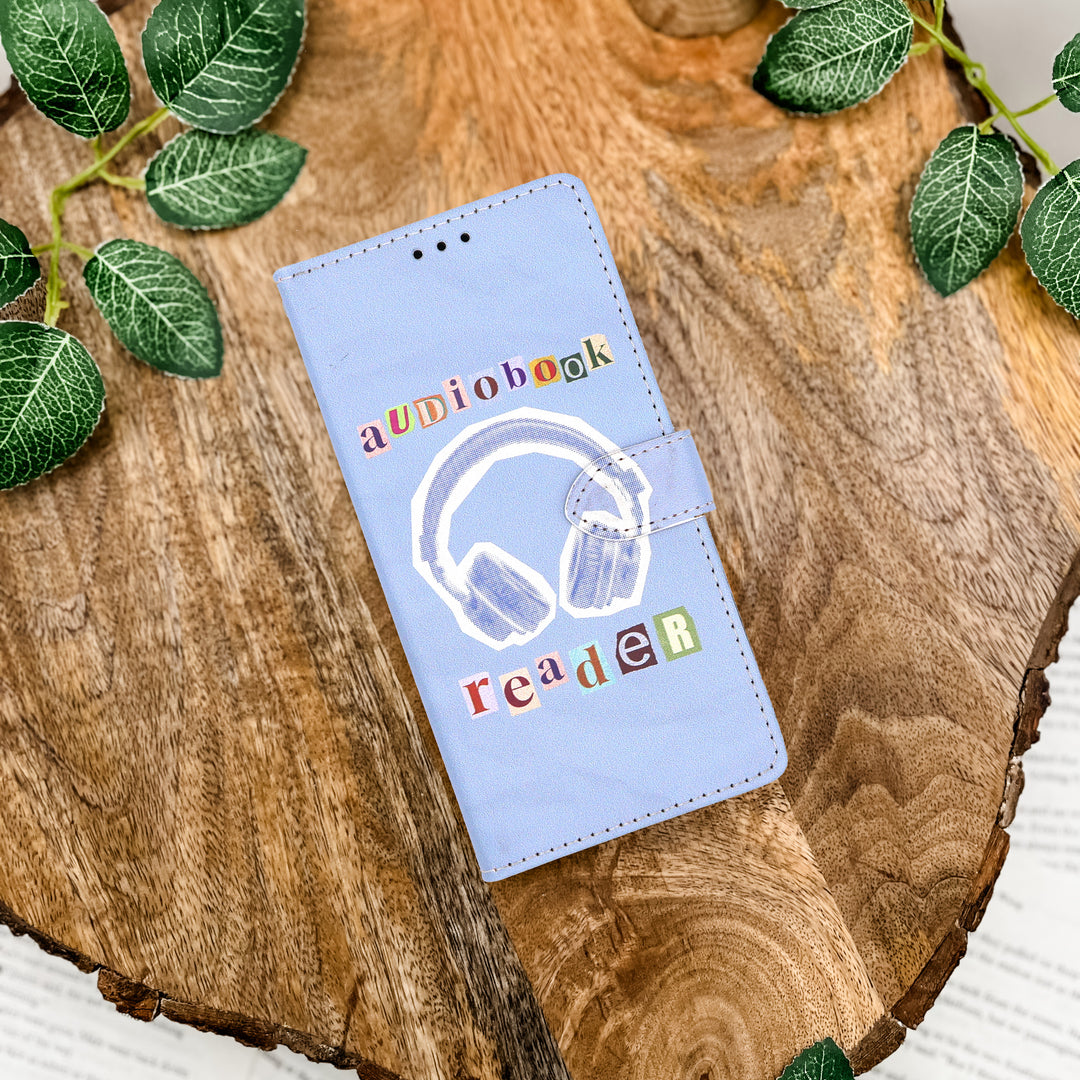 Light blue phone case with 'audiobook reader' text and headphones design on a wooden surface with green leaves.