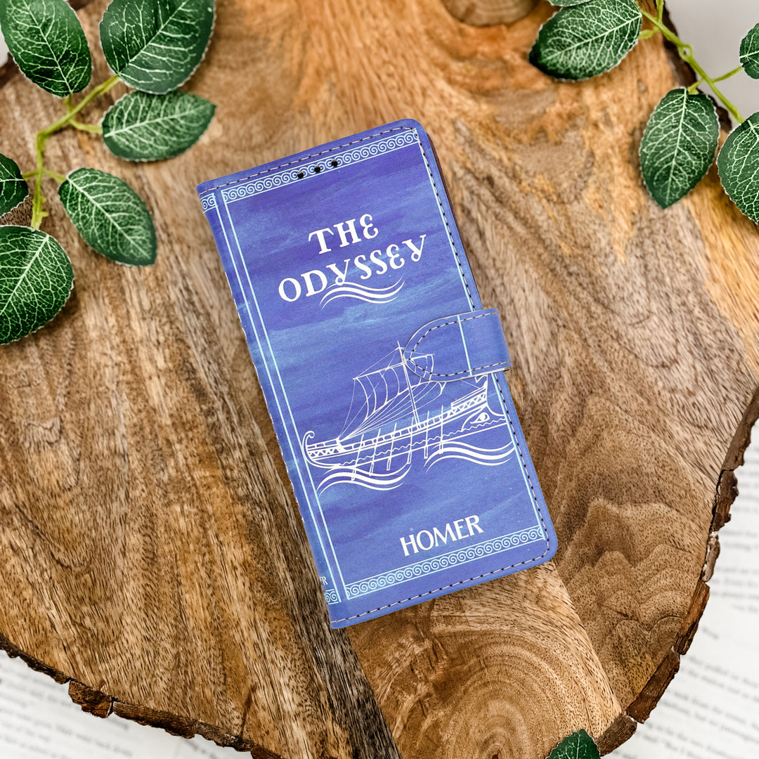 Blue book-shaped phone case with 'The Odyssey' design on a wooden surface with green leaves.