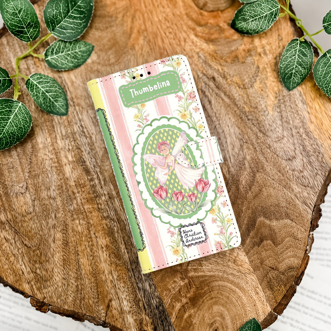 Book shaped phone case inspired by 'Thumbelina' with floral design on a wooden surface with green leaves.
