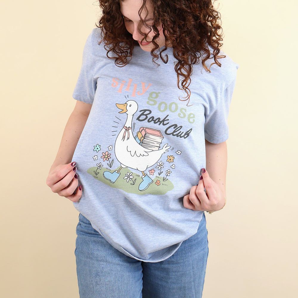 A woman wearing a light blue t-shirt with a Silly Goose Book Club graphic and text on a beige background