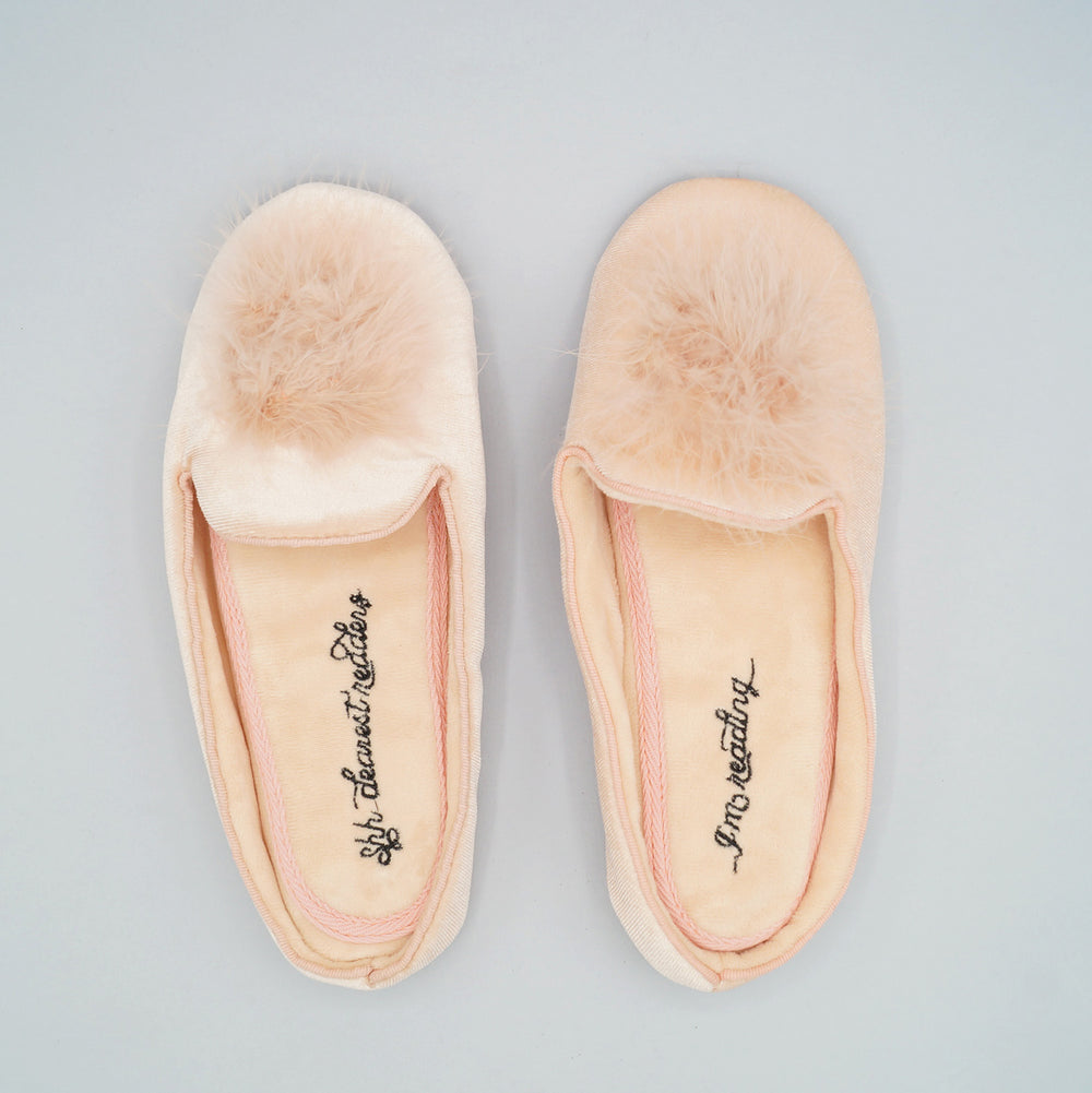 pale pink slip-on slippers with pompoms on the front. There is black writing embroidered on the inside bottoms of the slippers - the left says "Shh dearest readers" and the right says "...I'm reading..."