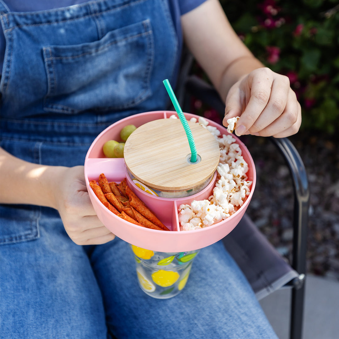Girl in blue overalls holds a cup with a light pink snack tray with four sections, each filled with snack food