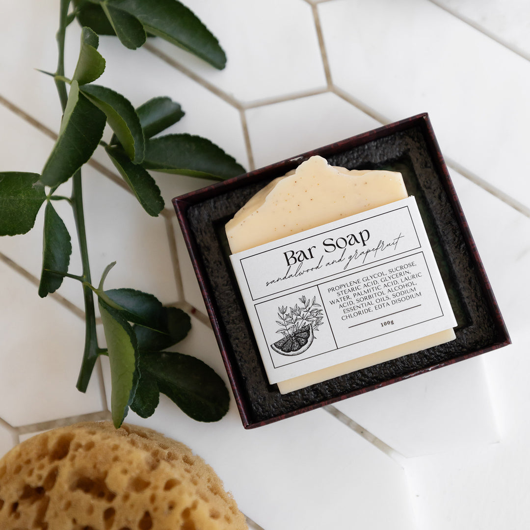 An open box with a bar of sandalwood and grapefruit soap next to a leaf.