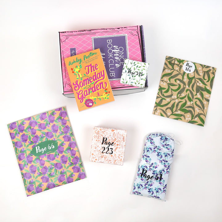 A paperback edition of The Someday Garden by Ashley Poston lays on a pink OUABC box next to a themed wrapped gift labeled with page numbers. In front of the pink OUABC box are four more themed wrapped gifts labeled with page numbers.