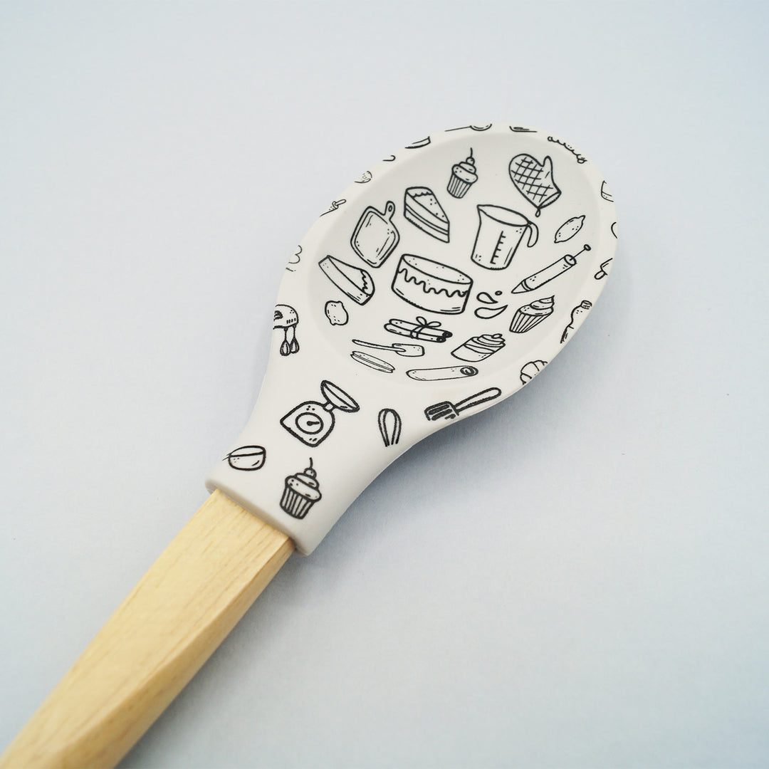 A white silicone cooking spoon with a wooden handle. There is a pattern of kitchen appliances of the silicone part.