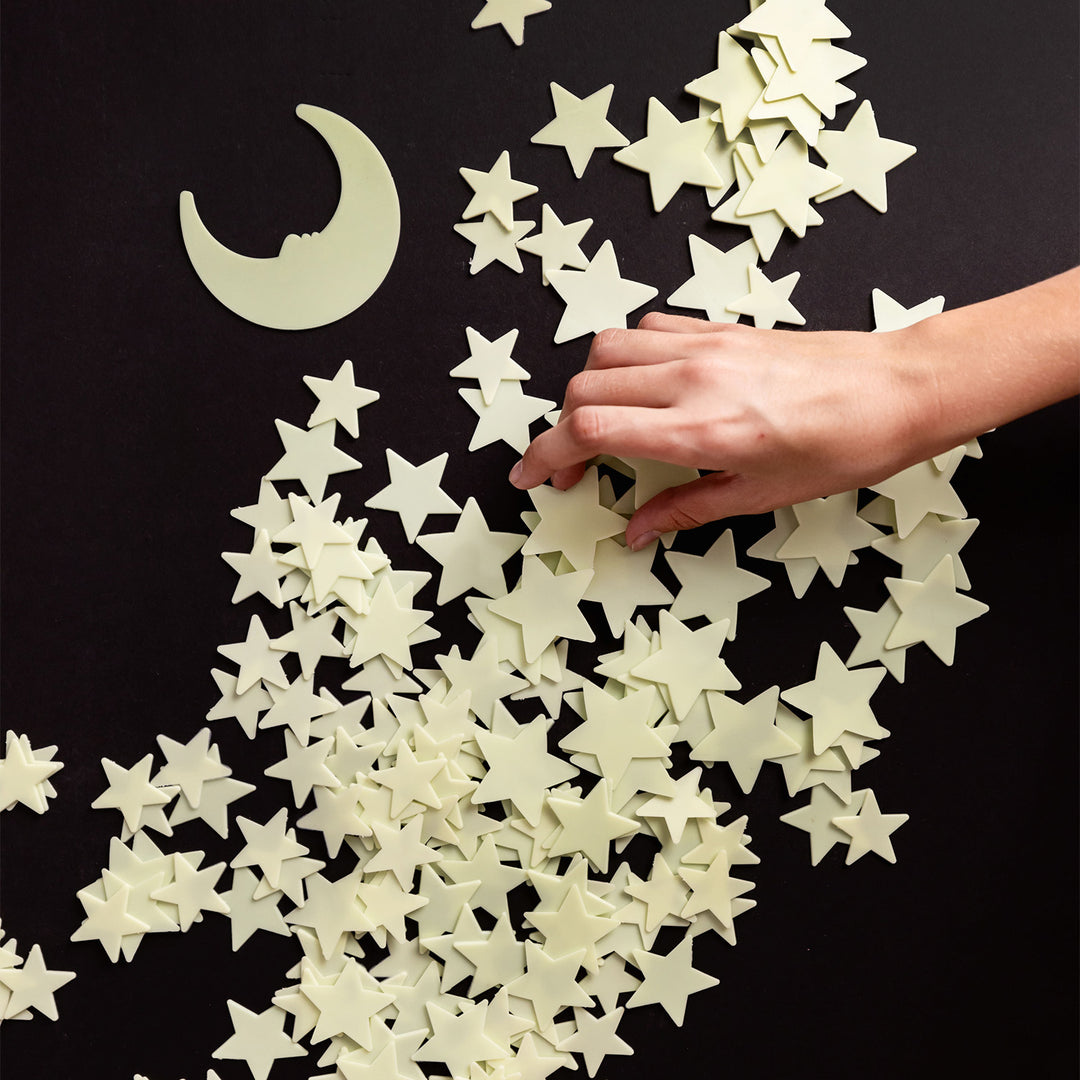 a pile of glow in the dark stars and a moon sit on a black background. A white hand is reaching to grab one from the center