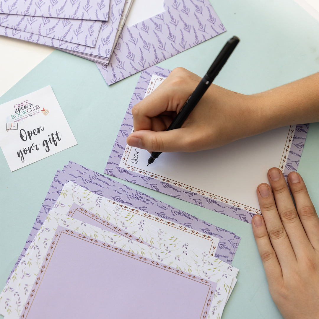 A hand holding a black pen writes on a purple page surrounded by the rest of the purple stationary set, next to an Open Your Gift sticker.