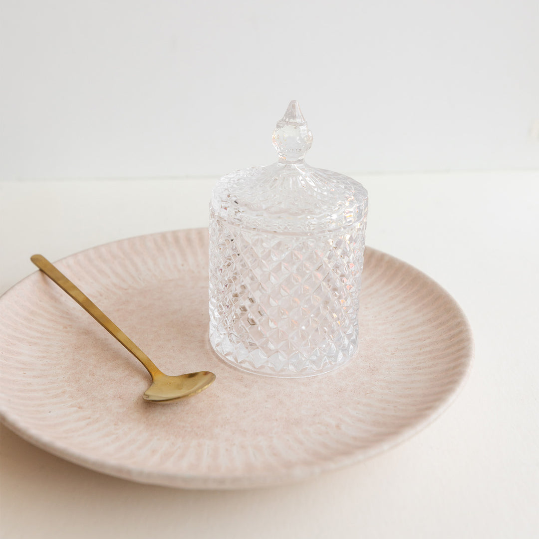 A crystal sugar jar with lid next to a long gold spoon