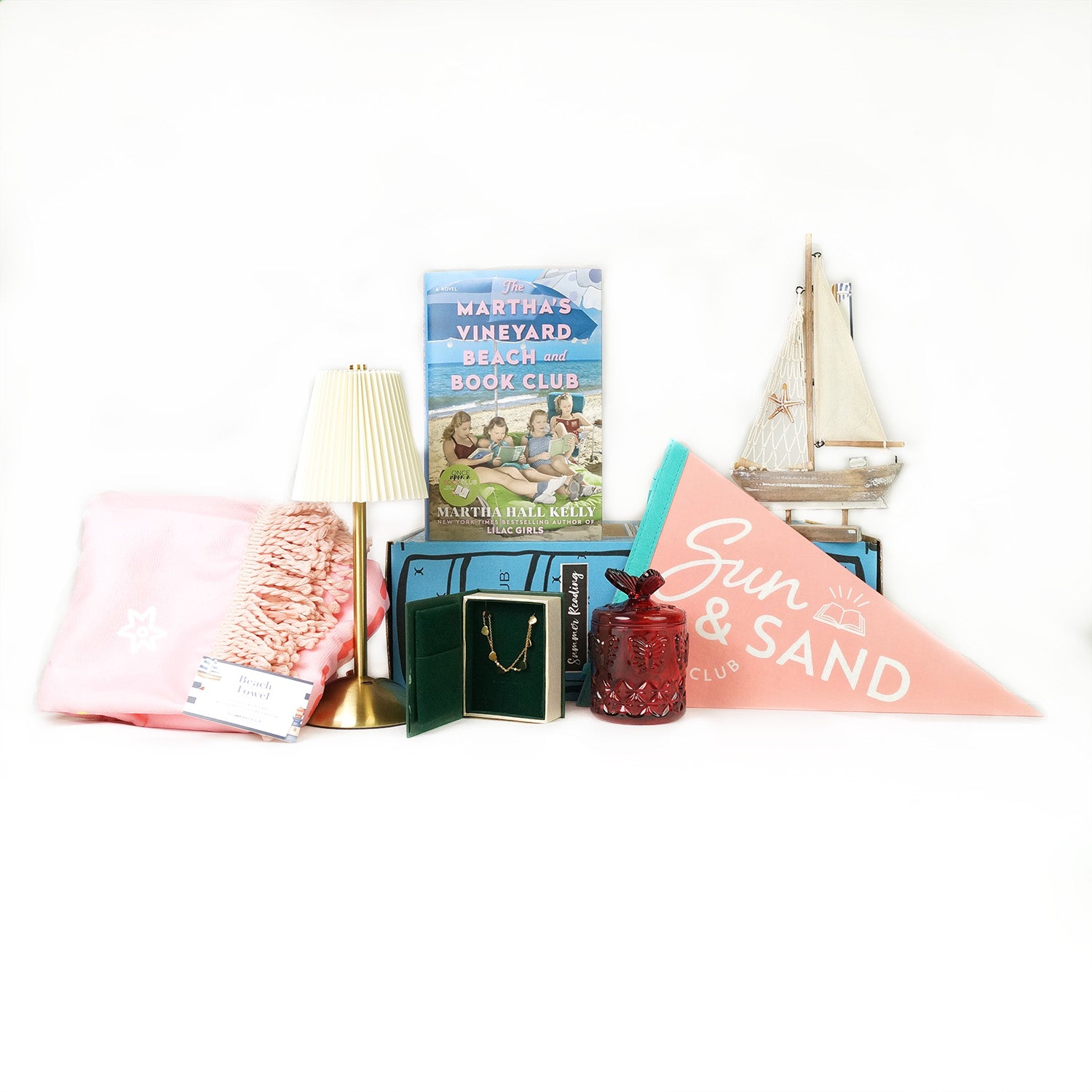 A hardcover edition of The Martha's Vineyard Beach and Book Club stands on a blue OUABC Summer Reading Box next to a wooden replica of a sailboat. In front of the box, from left to right, are a folded pink beach towel, a gold lamp with a white lampshade, an open box with a gold bracelet inside, a red glass jar with a butterfly on top, and a pink and teal pendant flag that says Sun & Sand Book Club on it.