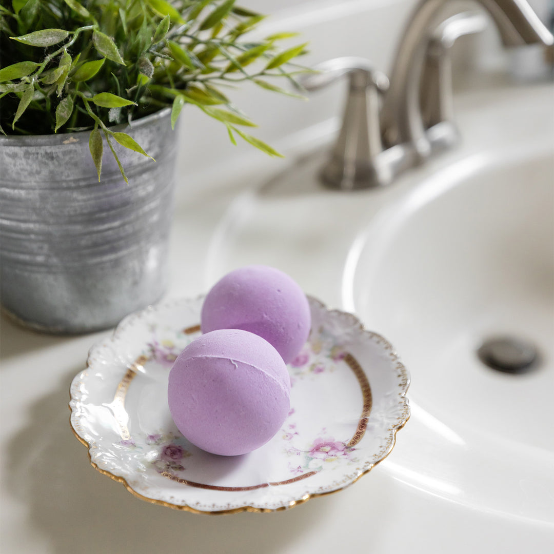 Two light purple bath bombs sit on an ornate plate next to the bathroom sink.