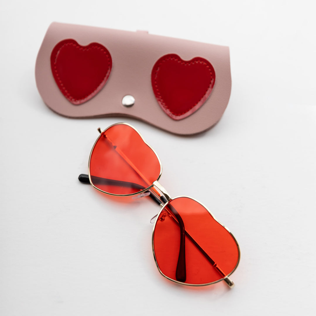 heart-shaped sunglasses with gold frames and red lenses next to a faux leather case with two red hearts on it