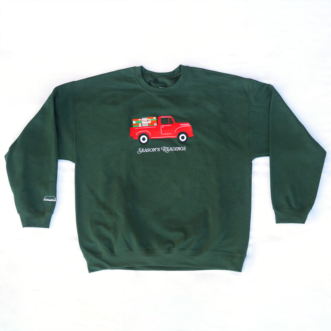 A forest green embroidered sweatshirt with a red pickup truck filled with books. Under the truck are the embroidered words "Season's Readings". On the right sleeve is a little white embroidered open book.