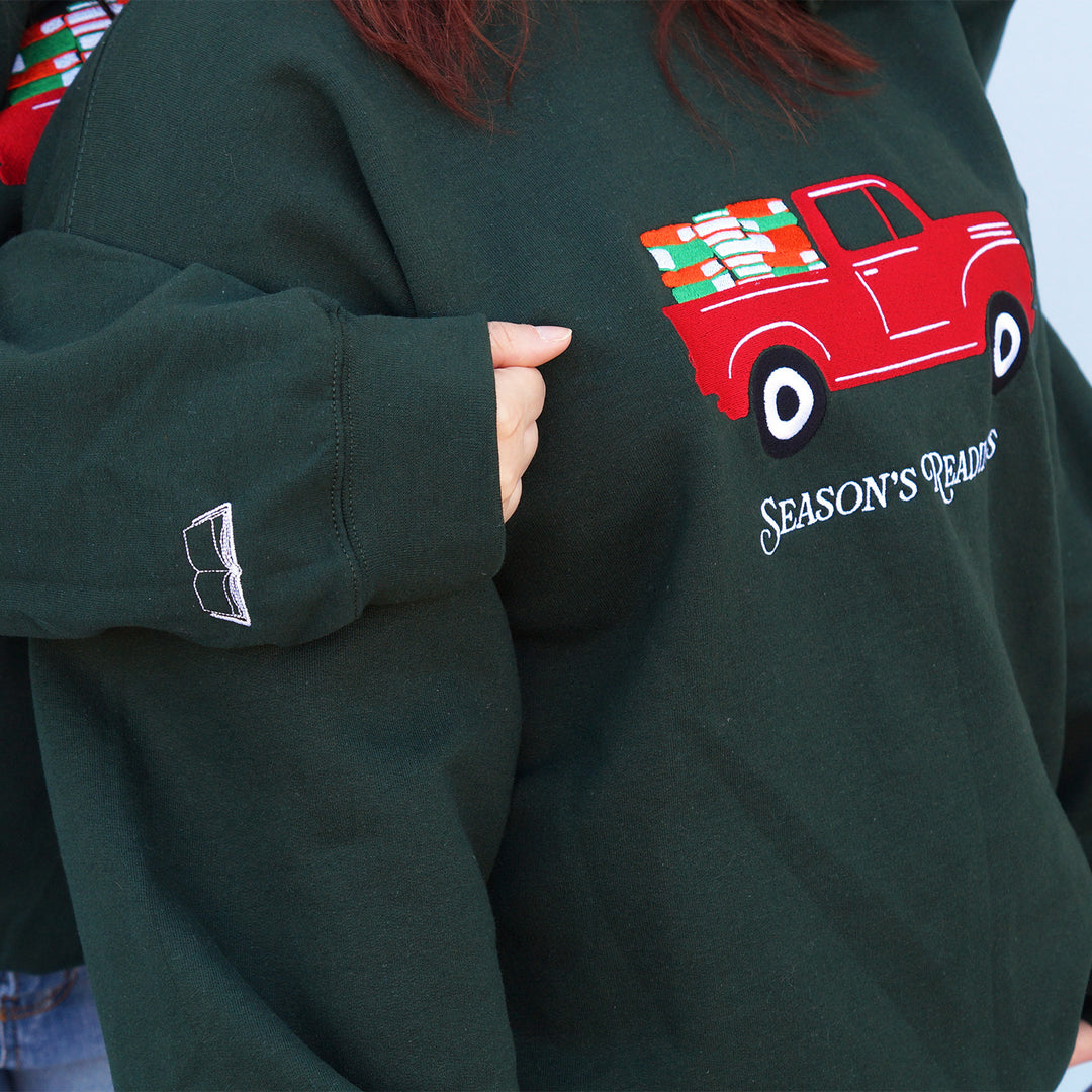 Green sweatshirt with an embroidered red truck graphic and 'Season's Readings' text on a person wearing it while another person has their arm on the first's shoulder showing off the embroidered arm detail.