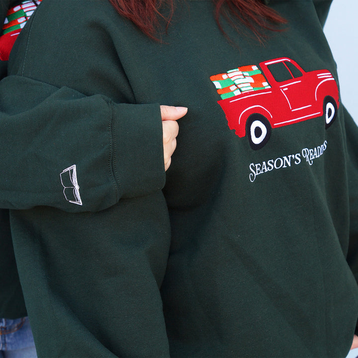 Green sweatshirt with an embroidered red truck graphic and 'Season's Readings' text on a person wearing it while another person has their arm on the first's shoulder showing off the embroidered arm detail.