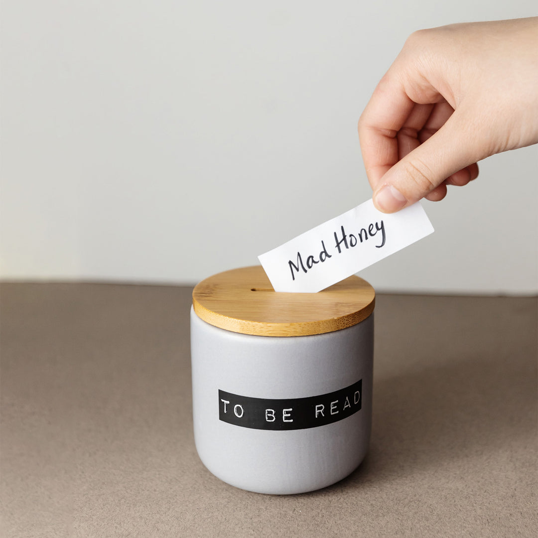 A gray, ceramic jar printed with black lettering reading 'TO BE READ' and a wooden lid. A white hand is putting a slip of paper that says Mad Honey in the opening at the top